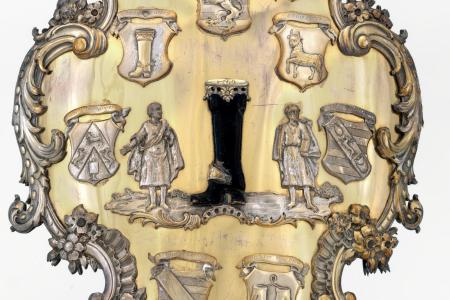 Groot schild van de Brugse schoenmakers en leerbewerkers  schild van zilver en koper  cartouchevormig borstschild in symmetrische Louis XV-stijl. Bovenaan in het middenveld het wapenschild van Brugge  in het centrum een gekroonde laars met datum '1569' tu