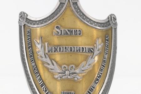 Schild van de Maatschappij Sint-Leopold te Brugge  met op de voorzijde een opschrift 'Sinte Leopoldus Maetschappy  der Vereenigde ambagtslieden 1939' en twee gekruiste Lauriertakken met strik binnen en omlijsting van palmetten. De bekroning is een mijter