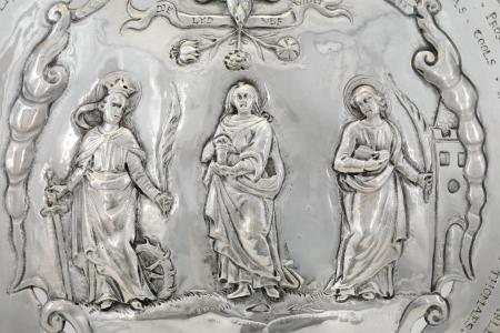 Schild van zilver  voorstelling van de heiligen Catharina  Magdalena en Barbara met bovenaan een duif met drie bloemen in de bek