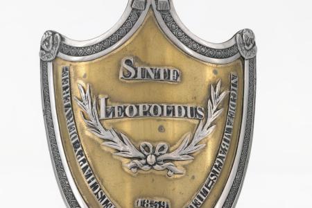 Schild van de Maatschappij Sint-Leopold te Brugge  met op de voorzijde een opschrift 'Sinte Leopoldus Maetschappy  der Vereenigde ambagtslieden 1939' en twee gekruiste Lauriertakken met strik binnen en omlijsting van palmetten. De bekroning is een mijter