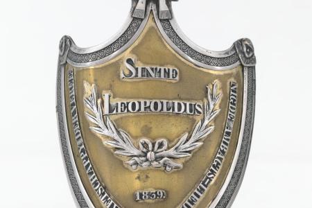 Schild van de Maatschappij Sint-Leopold te Brugge  met op de voorzijde een opschrift 'Sinte Leopoldus Maetschappy  der Vereenigde ambagtslieden 1939' en twee gekruiste Lauriertakken met strik binnen en omlijsting van palmetten. De bekroning is een mijter